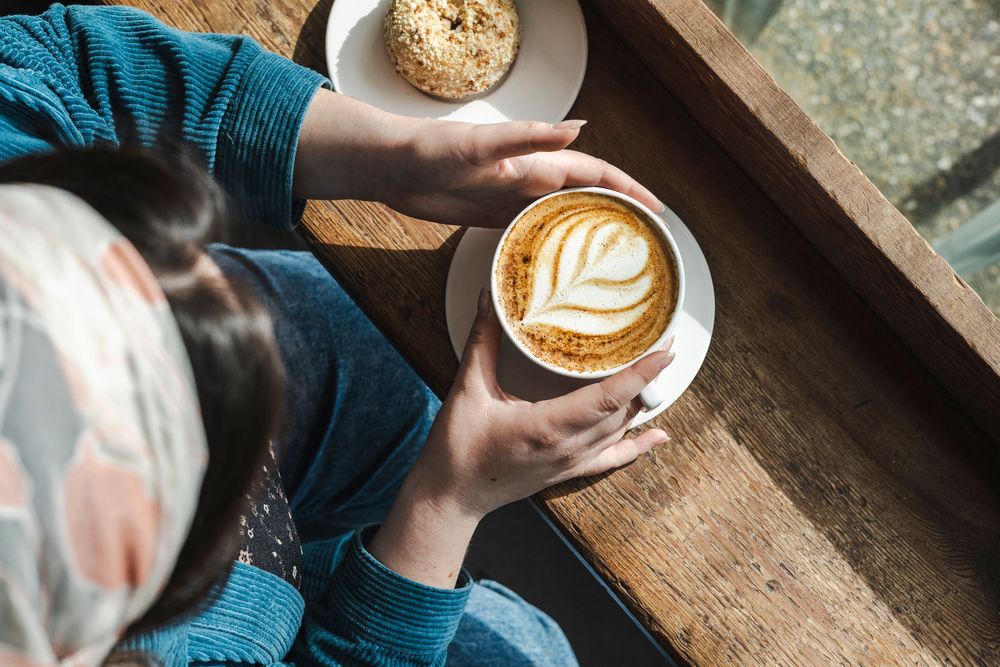 Hands holding a latte with leaf art design and a cookie on rustic wooden table.