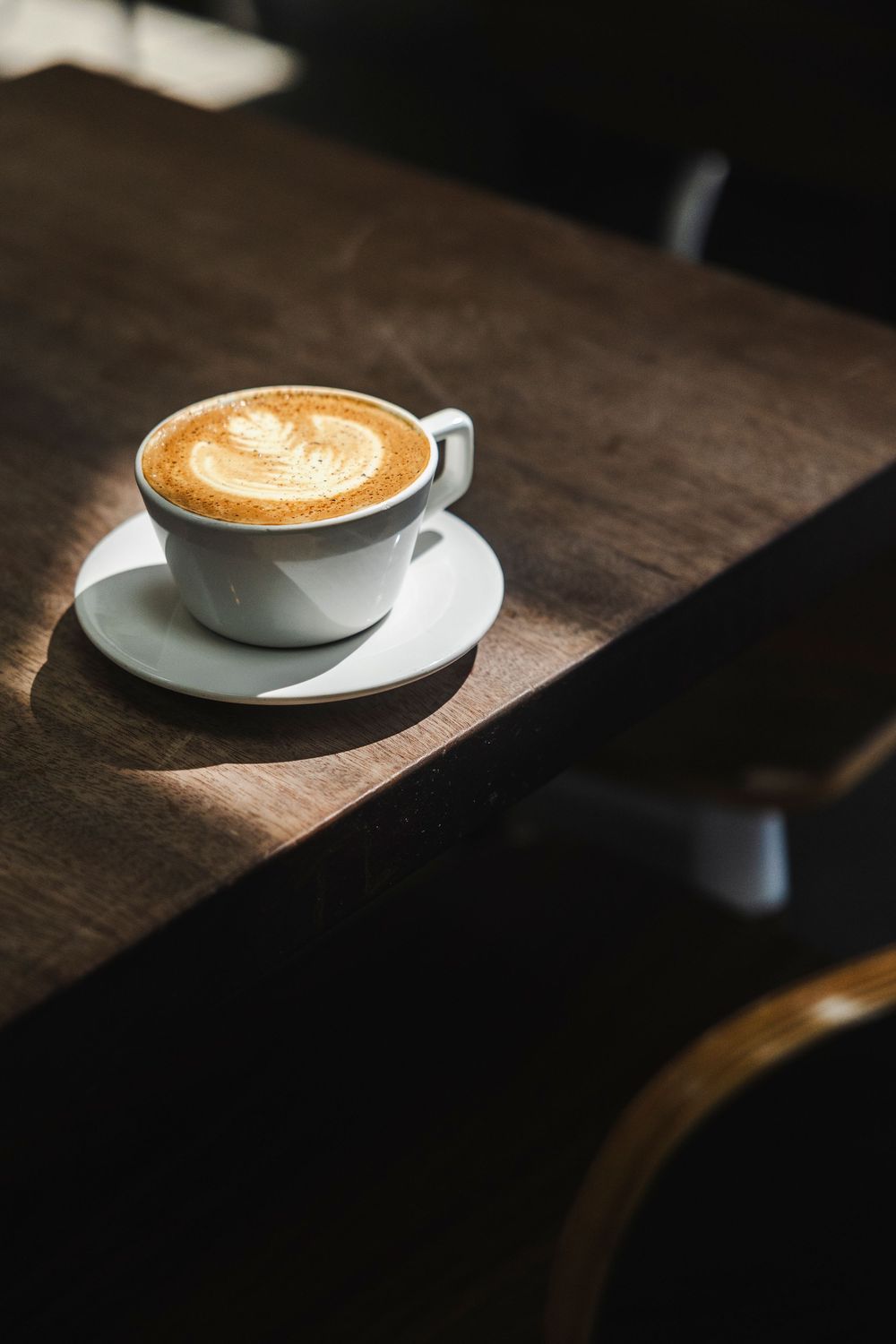 White coffee cup with latte art sitting on a dark wooden table in dramatic lighting.