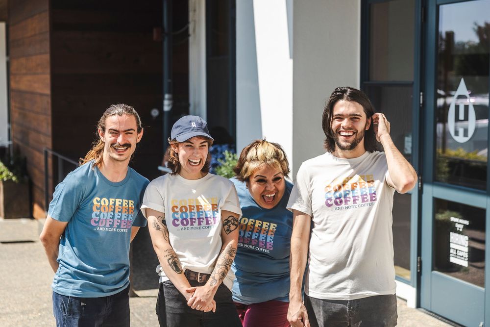 Four people wearing different colored Subtle Alchemy branded t-shirts smile together outside a storefront on a sunny day.