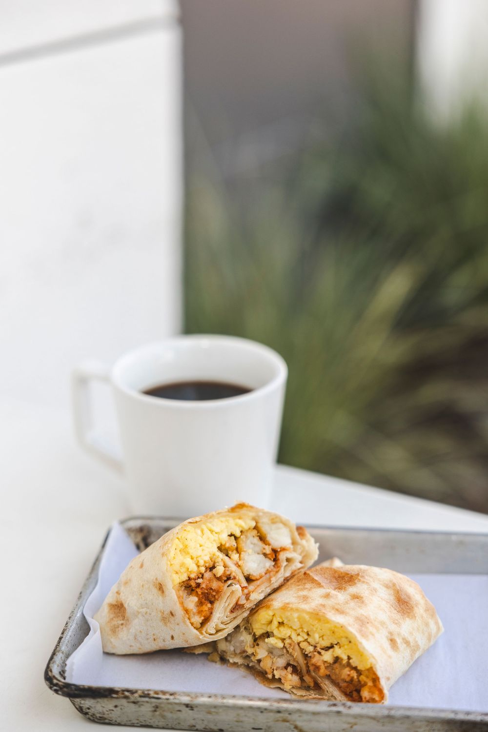 A breakfast burrito cut in half served on a metal tray with a white coffee cup in the background.