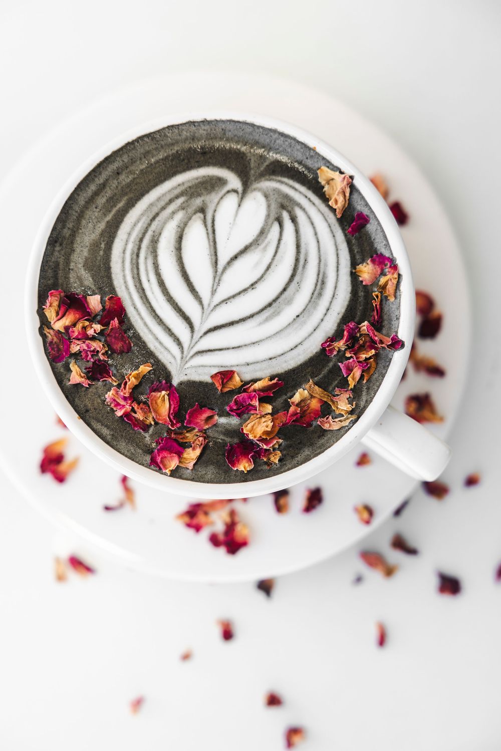Latte art design with a heart pattern surrounded by dried rose petals in a dark ceramic cup.