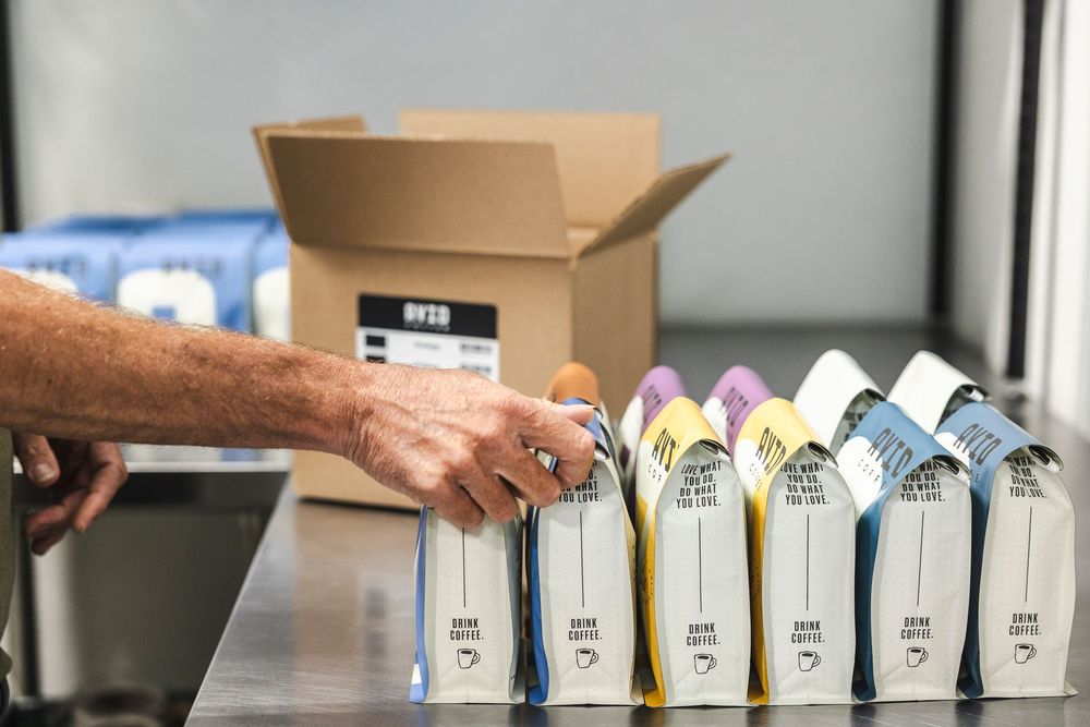 A hand arranges rows of colorful drink bottles near a cardboard shipping box on a shelf.