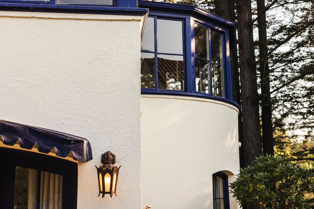 A cozy white lighthouse with blue trim and a lantern fixture stands among evergreen trees at sunset.