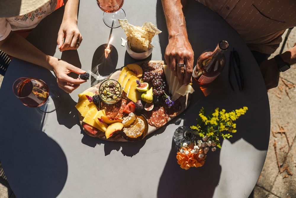 Overhead view of people sharing a charcuterie board with wine and yellow flowers on an outdoor table in sunlight.