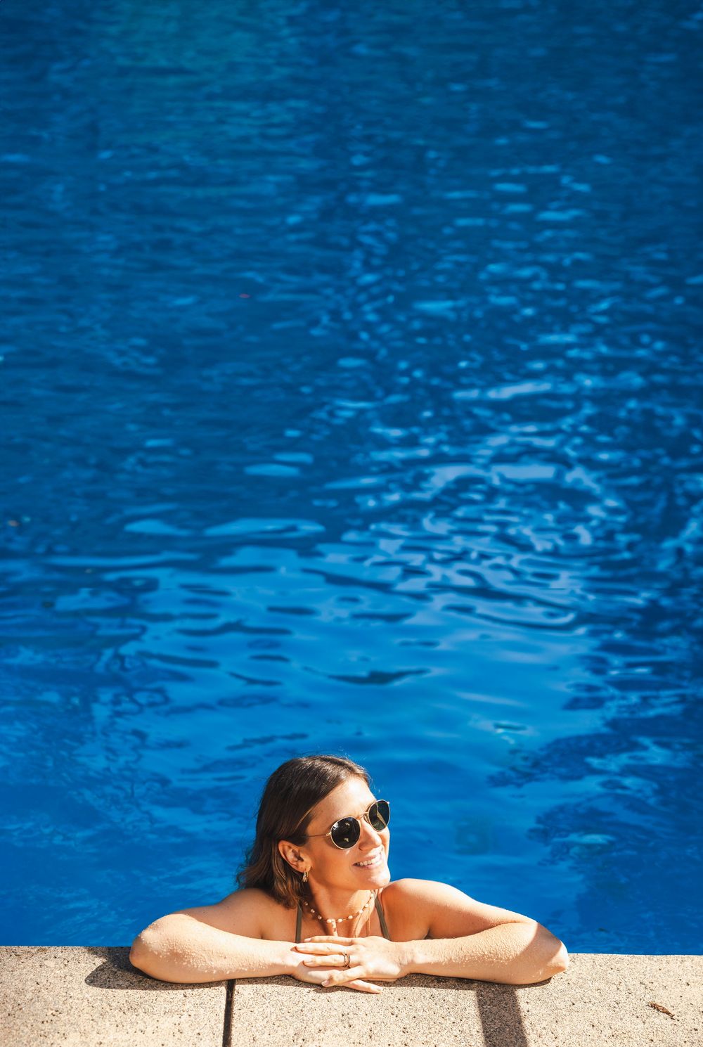 Person relaxing at edge of bright blue swimming pool wearing sunglasses on sunny summer day.