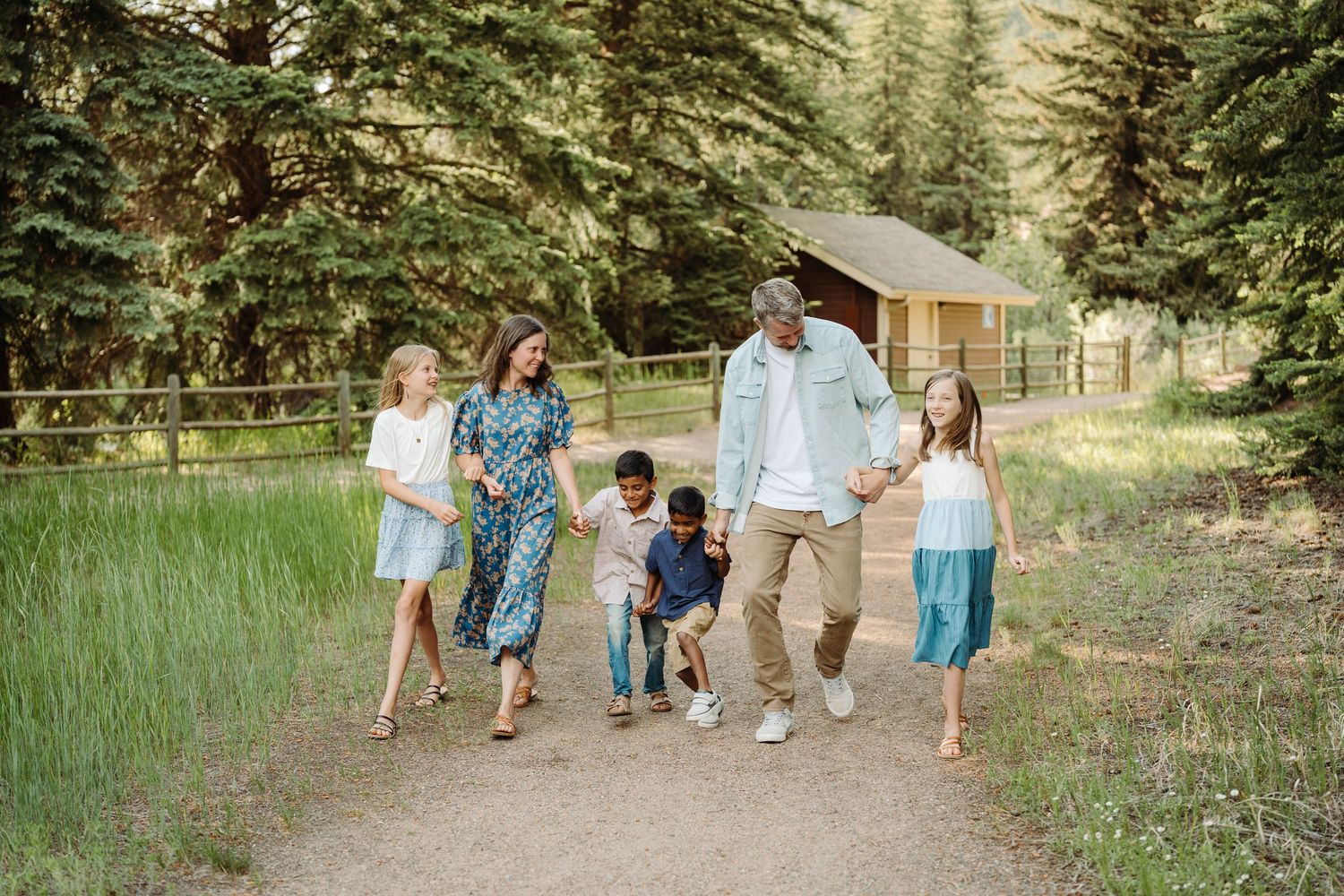 A family walks together on a dirt path through green grass and pine trees with a rustic cabin in the background.