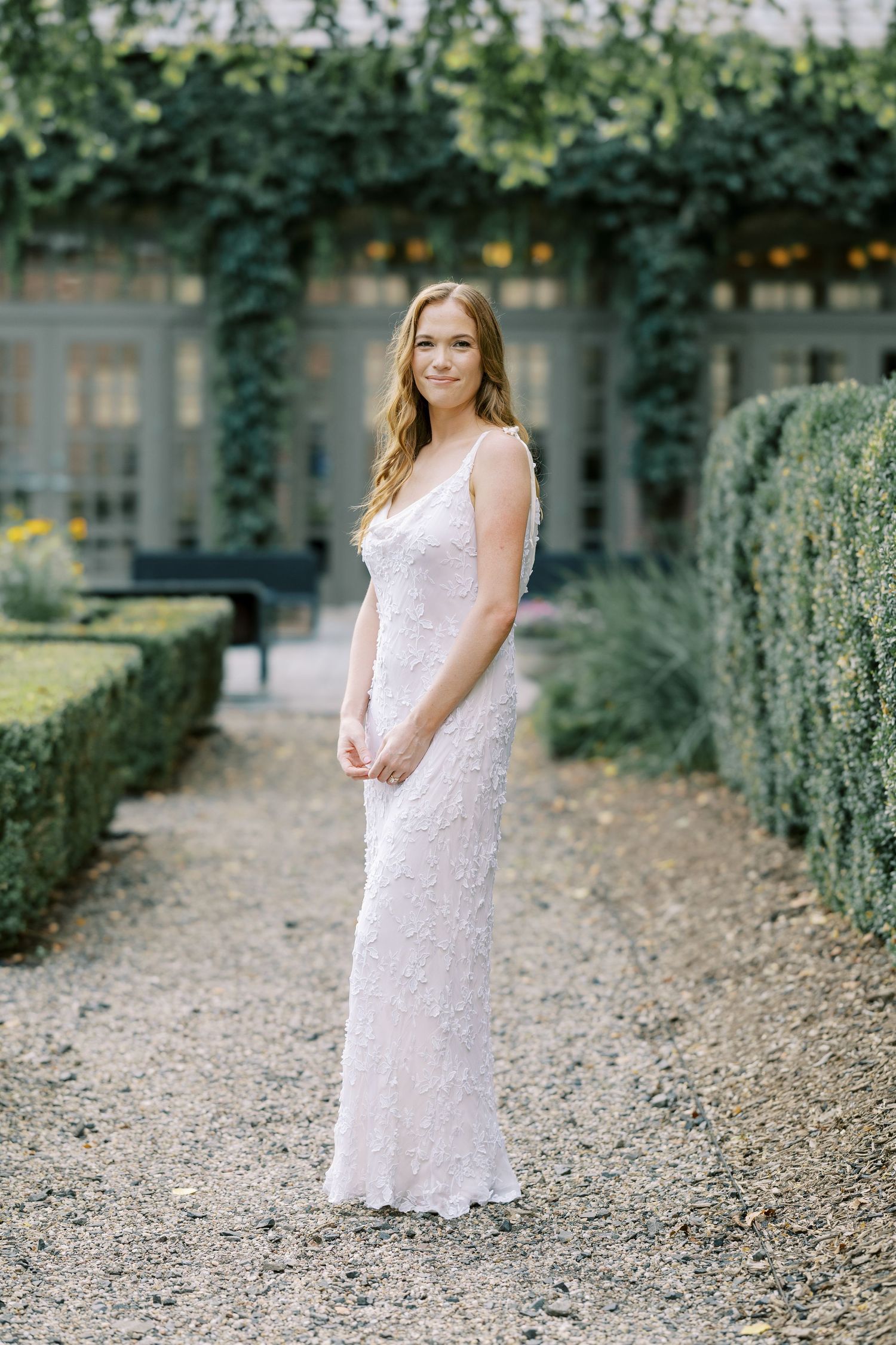 Bride in a white lace dress poses on a garden path lined with manicured hedges and trees on a sunny day at Ninety Acres.