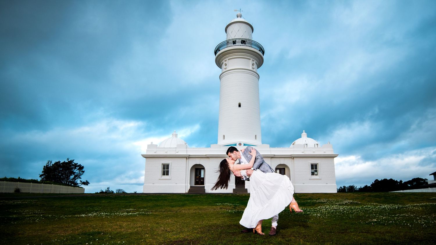 A lighthouse stands tall against a dramatic cloudy sky on a grassy hilltop near the ocean, where a couple is kissing on their wedding day.