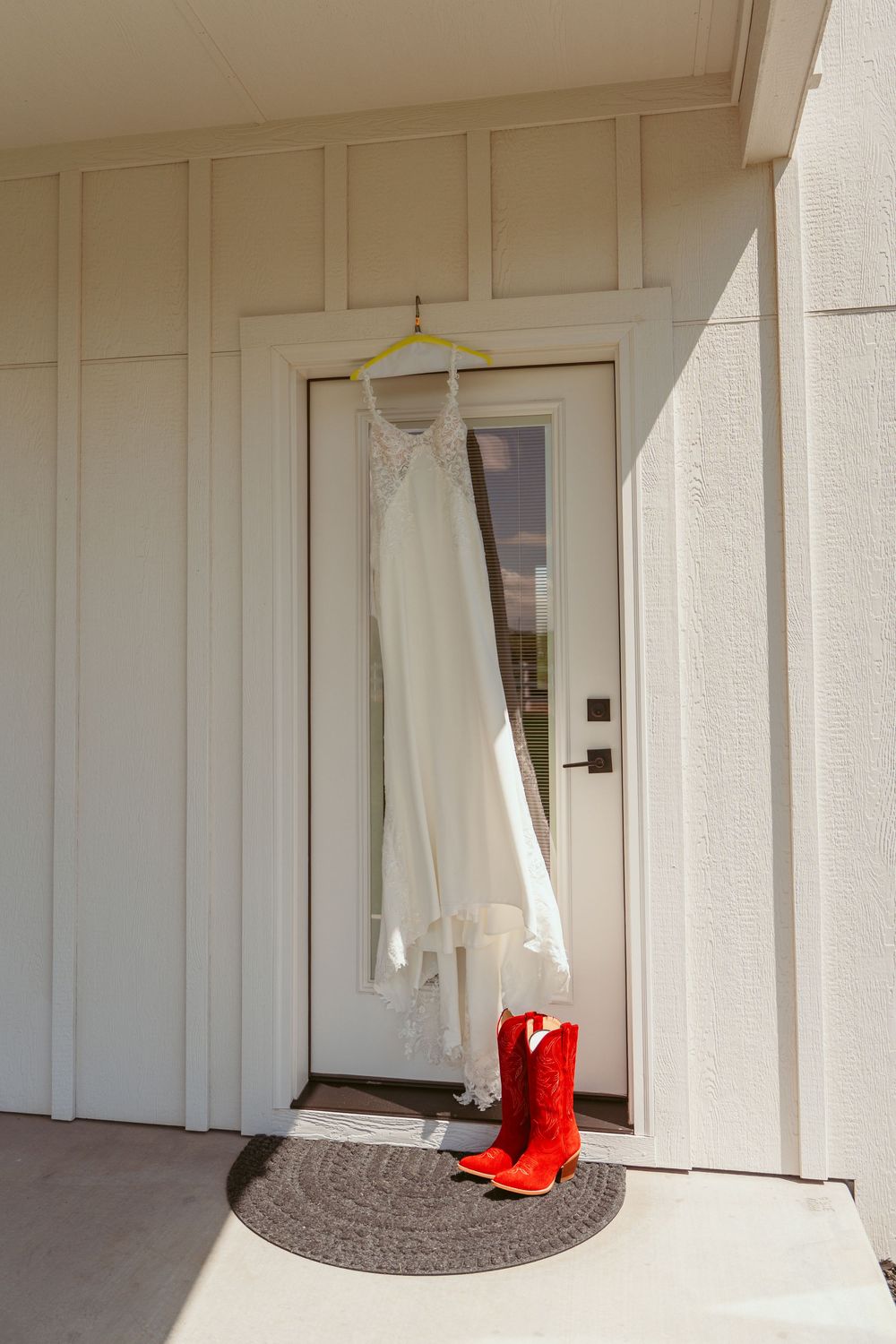 A white wedding dress hangs on a hanger outside a white door with bright red cowboy boots placed below on a doormat.