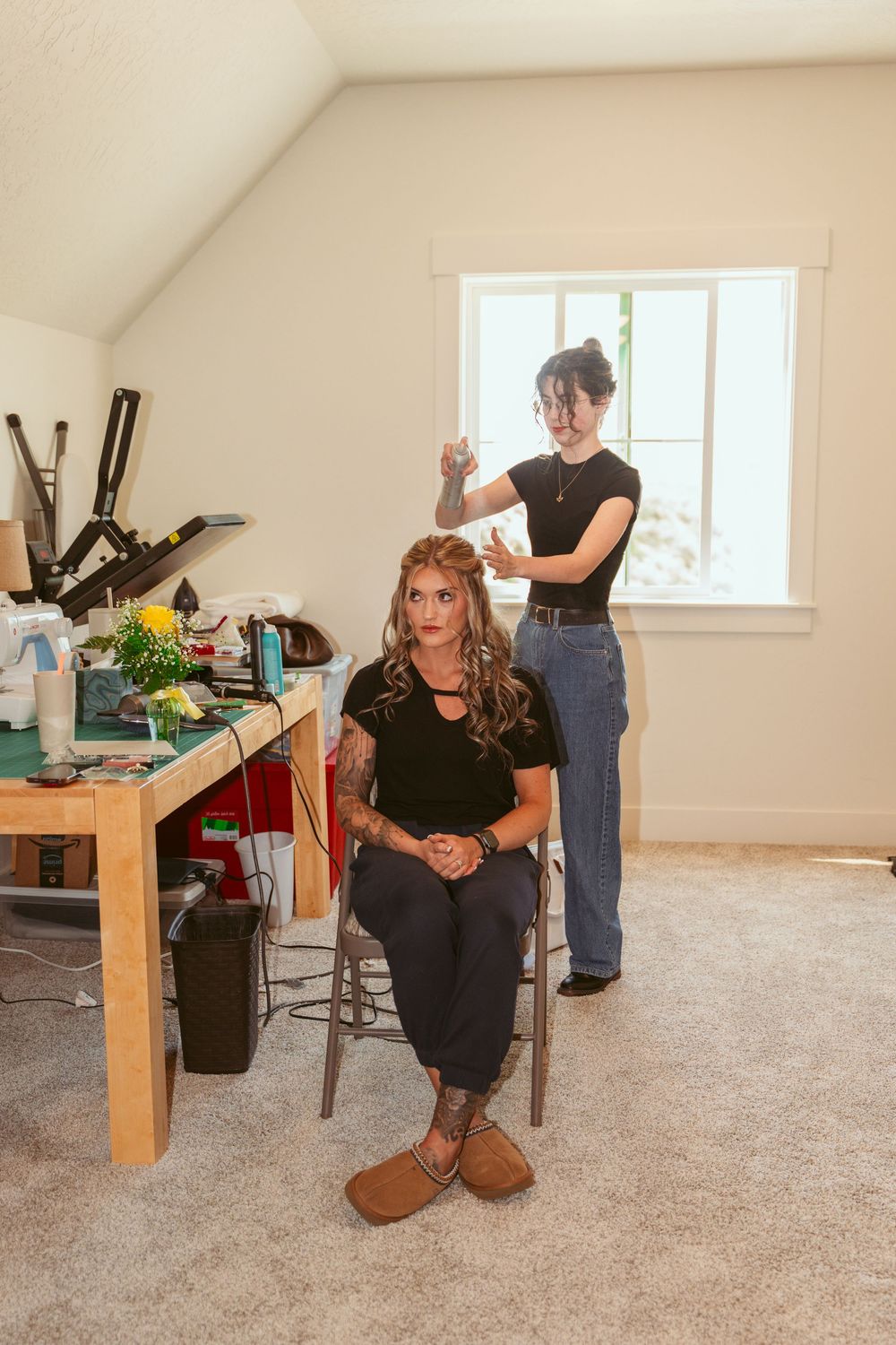 A hairstylist styling hair in a bright room with a wooden table and carpeted floor.