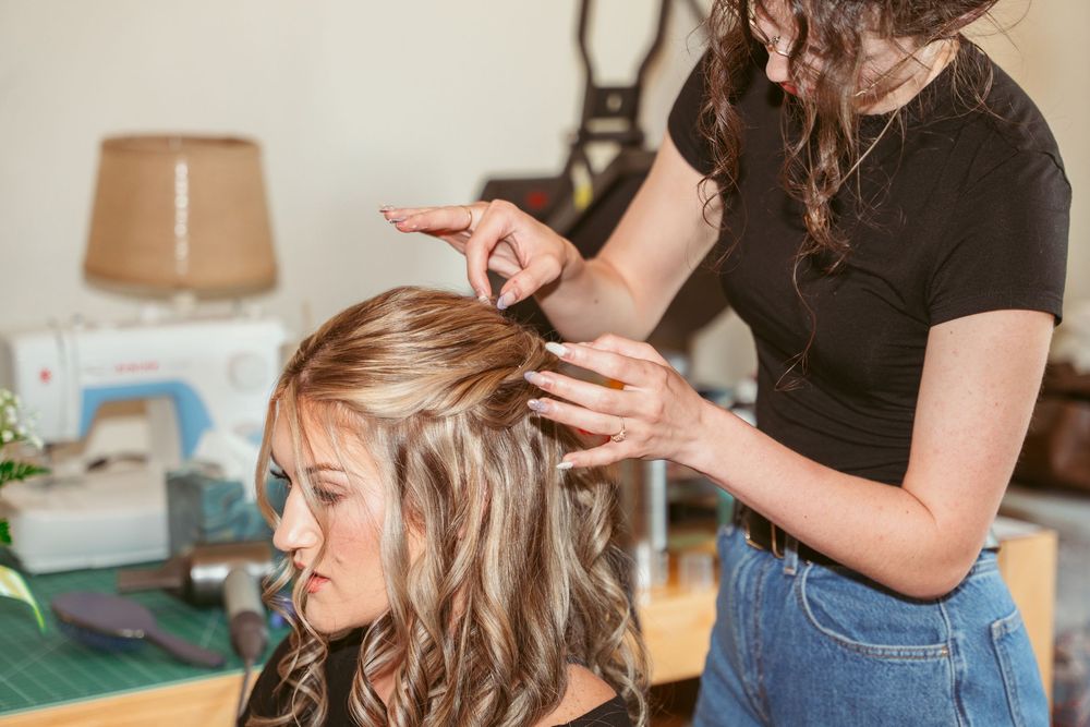 A hairstylist works on styling and curling long blonde hair in a home setting with a lamp in the background.