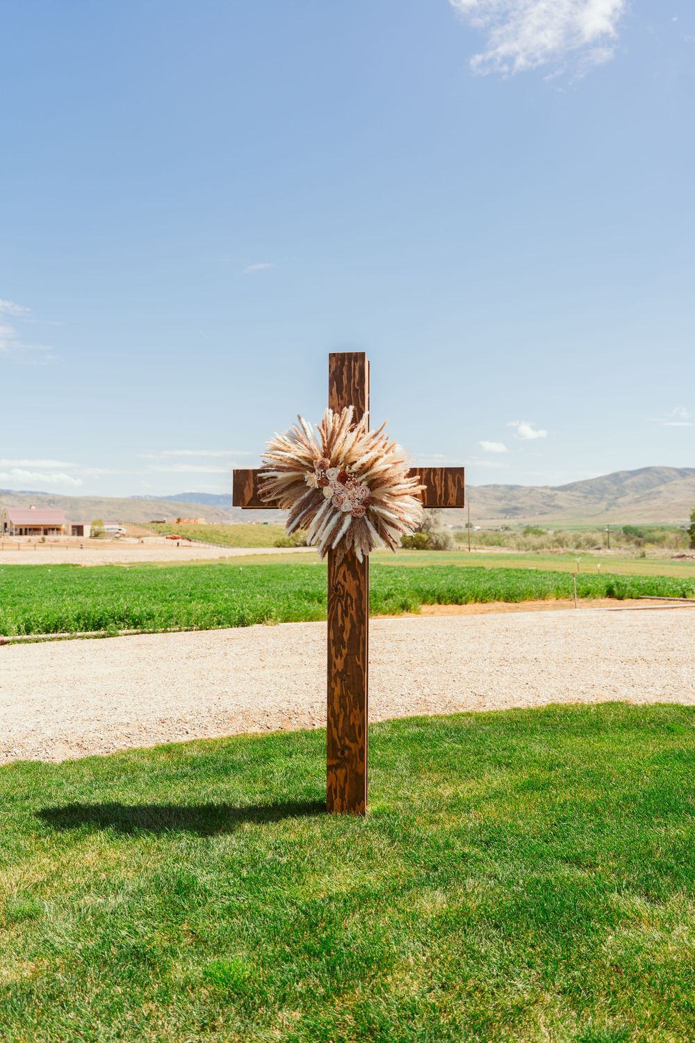 A wooden cross decorated with dried flowers stands on green grass against a mountain landscape.