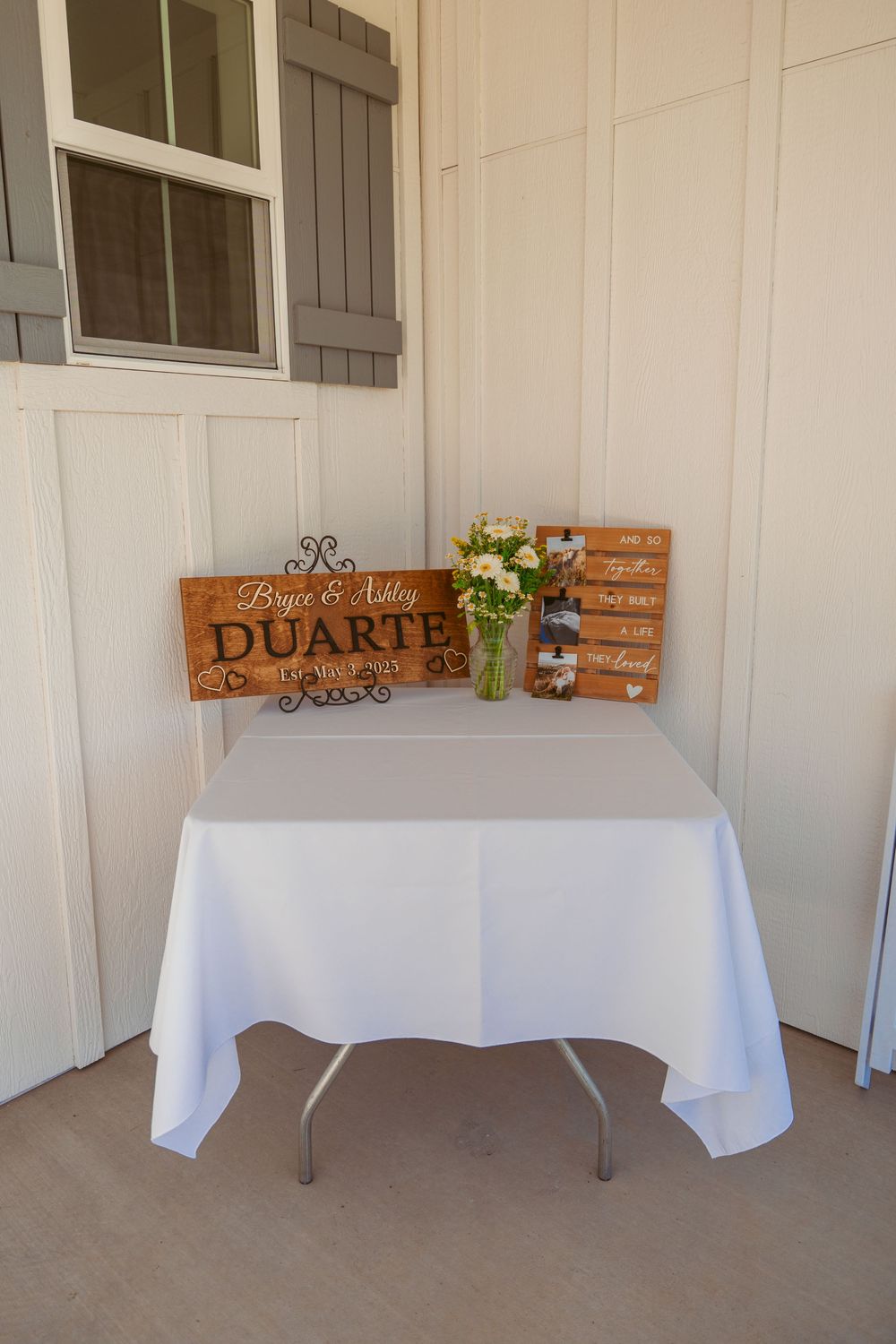 White-clothed reception tables with rustic wooden name signs and decor against a light-colored wall.