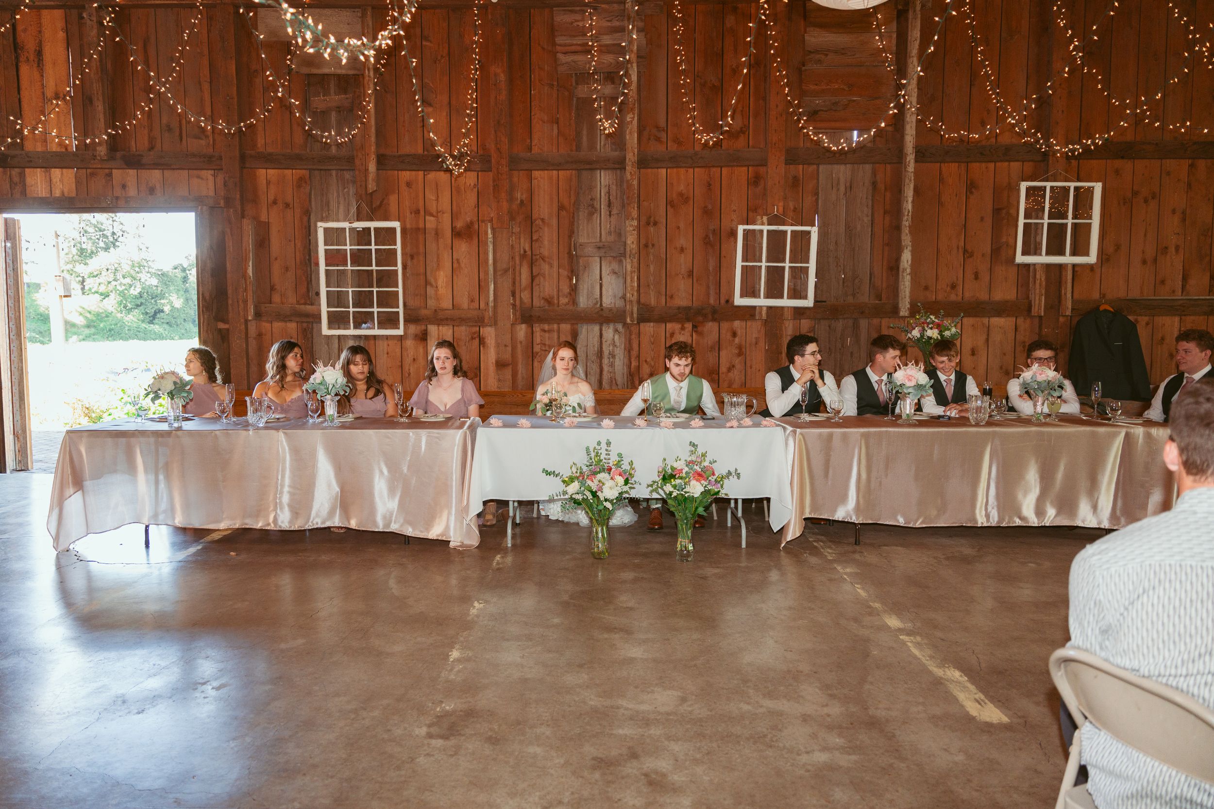 Long head table setup inside a rustic wooden barn with string lights decorating the ceiling.