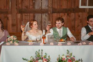 Wedding guests are seated at long tables decorated with floral centerpieces inside a rustic barn venue.
