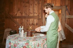 Couple cutting wedding cake at reception with floral tablecloth in rustic barn setting.