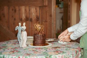 A chocolate wedding cake sits on a floral tablecloth as someone cuts into it for serving.