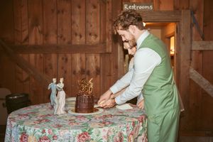 A person in a green vest cuts into a cake on a floral tablecloth with decorative figurines on top.