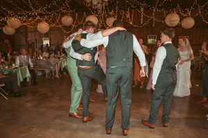 Wedding party members dancing together in a rustic barn with twinkling lights and paper lanterns overhead.
