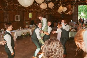 Wedding party members dancing and celebrating in a barn venue with twinkling lights and white decor.