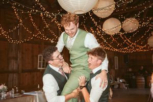 Joyful celebration with groomsmen lifting someone during a wedding reception in a decorated barn venue.