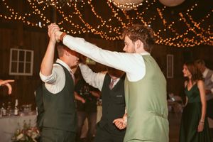 Wedding party members dance energetically under decorative string lights at a barn reception.