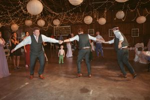 Groomsmen perform synchronized dance moves at barn wedding reception under string lights.