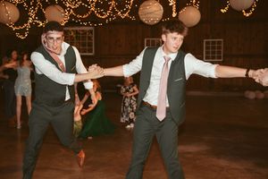 Two groomsmen dance energetically on the reception floor under decorative lights and lanterns.