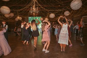 Wedding guests dancing and celebrating on the dance floor under string lights and paper lanterns in a rustic barn venue.