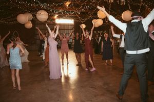 Wedding guests dancing and celebrating at a barn reception with festive lighting and paper lanterns overhead.