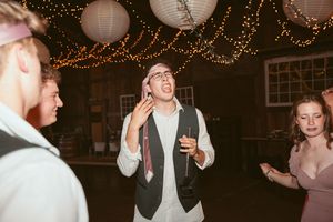 Wedding party members celebrating with drinks and laughter under decorative string lights at a barn reception.