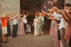 Wedding guests hold sparklers creating a glowing archway outdoors at night in front of a rustic barn.