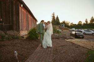 A couple walks hand in hand on a path outside a rustic farm building during sunset.