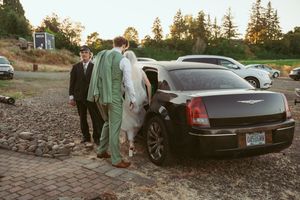 Wedding party members in formal attire gather beside a luxury car in an outdoor parking area at sunset.