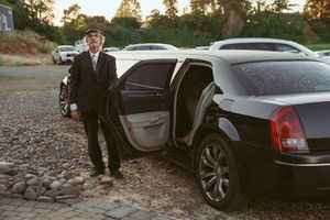 A well-dressed person stands beside a luxury black car with an open door on a gravel surface at dusk.