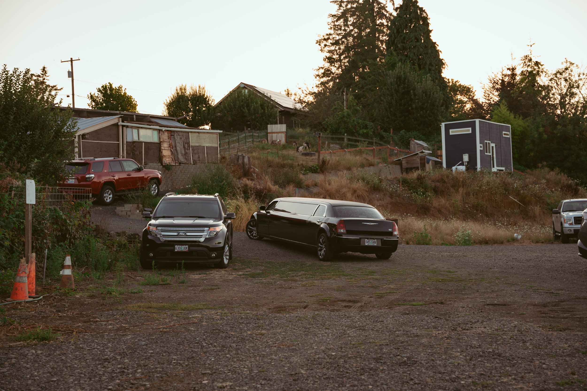 Cars parked in a gravel lot at dusk with residential buildings and trees visible in the background.