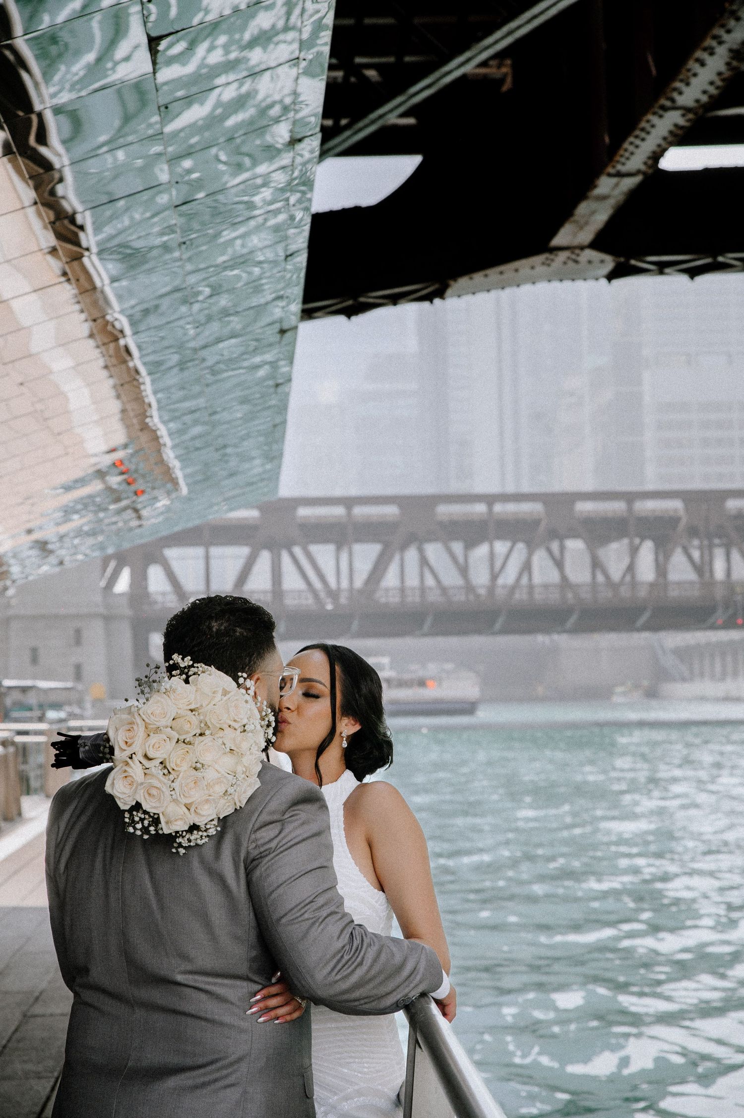 Couple embraces under bridge in Chicago with teal river water and industrial architecture in background on overcast day.
