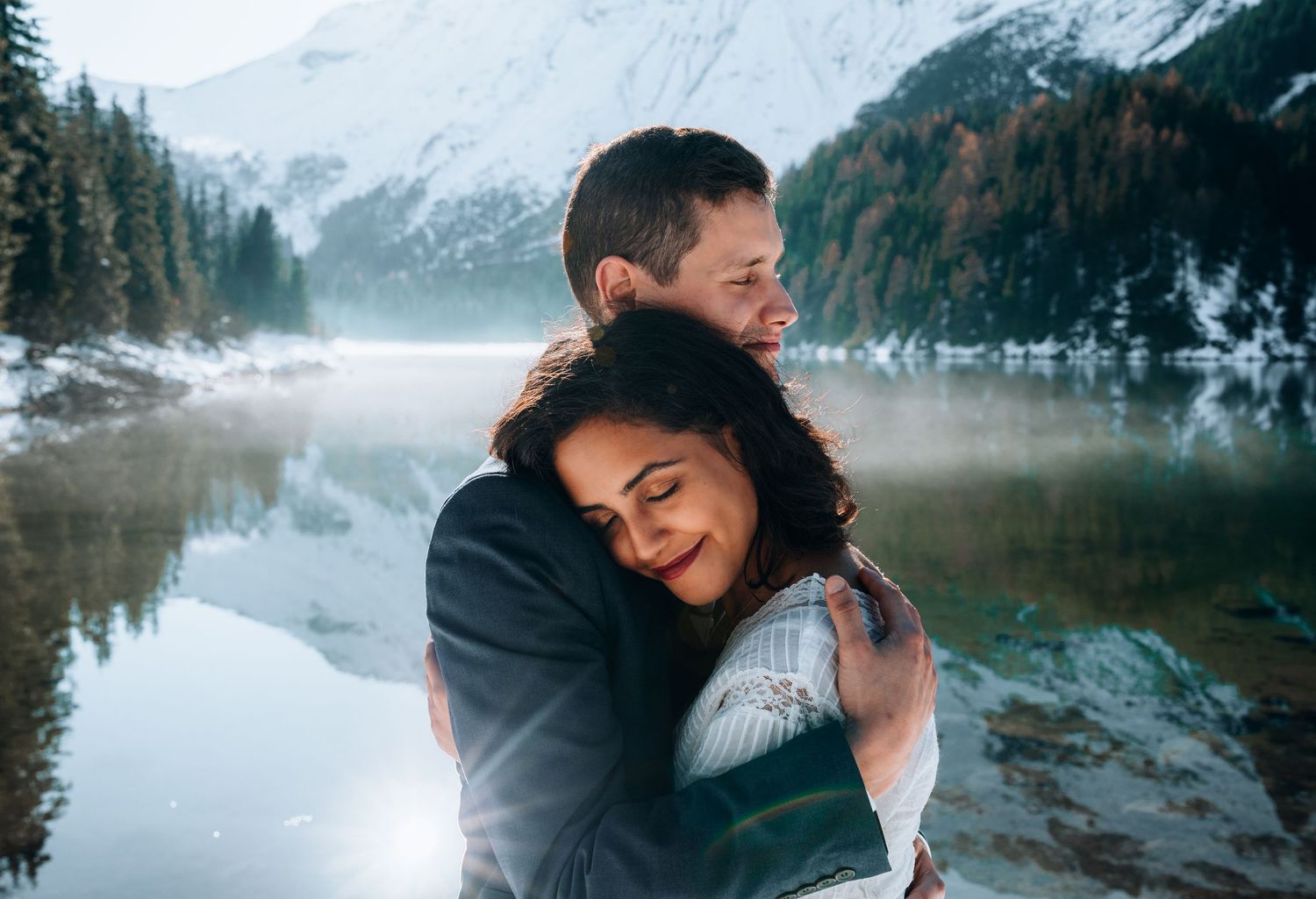 Two people share an intimate moment along a misty mountain lake with snow-capped peaks in the background.