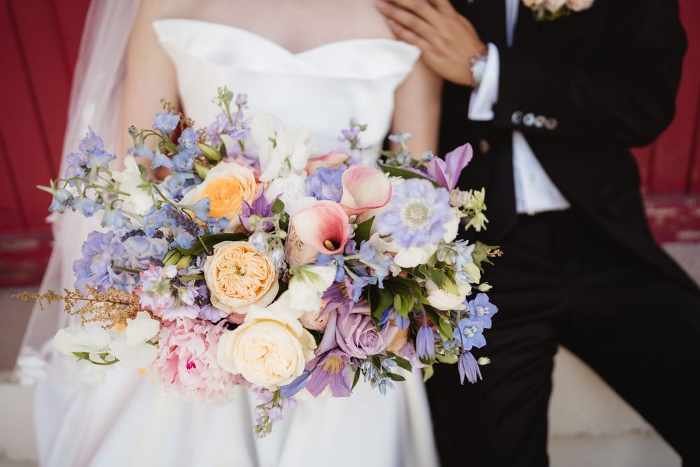 A pastel floral bouquet features peach garden roses and purple delphinium arranged naturally for a wedding.