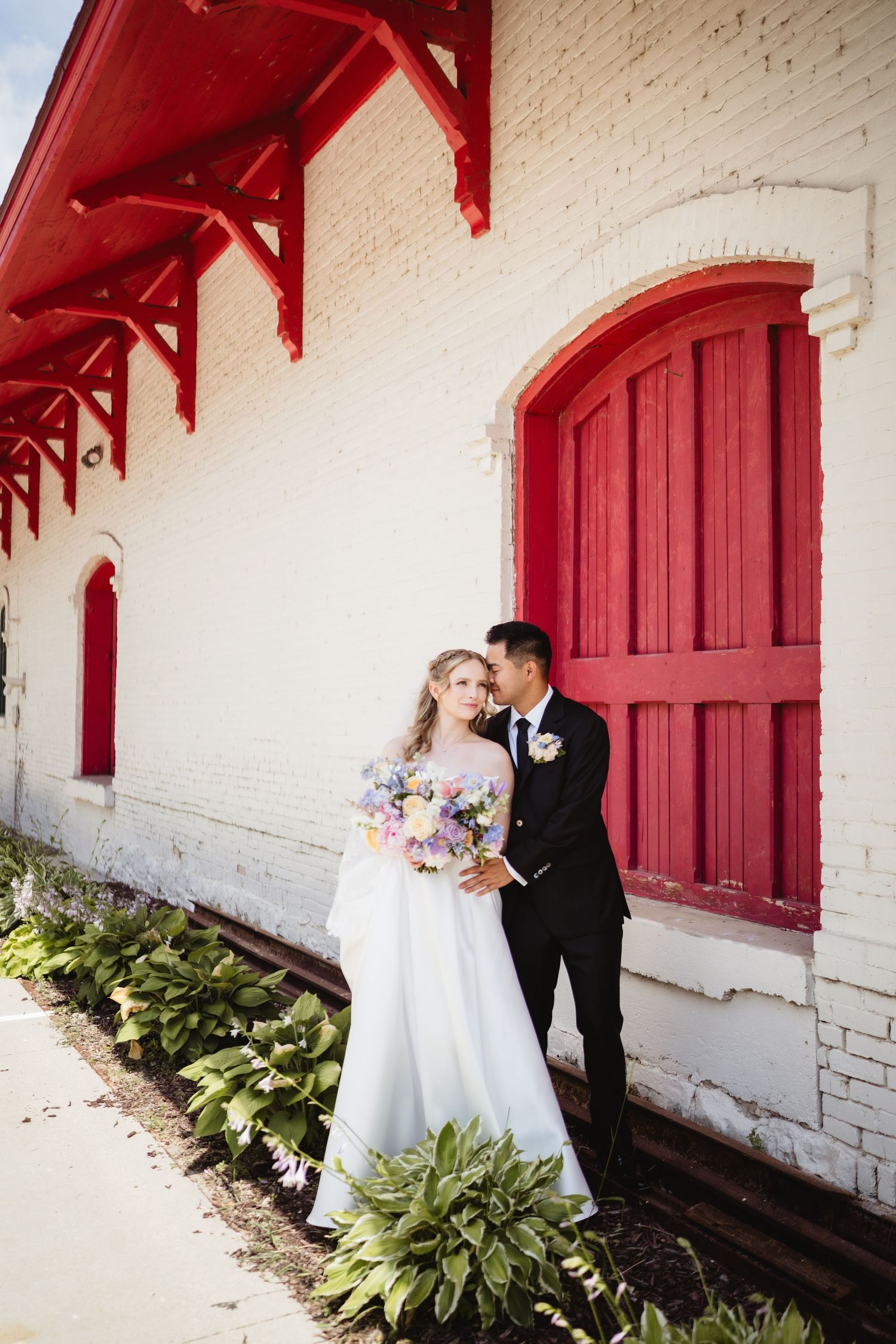 A couple in wedding attire poses for romantic photos against a white wall with red doors and green foliage.