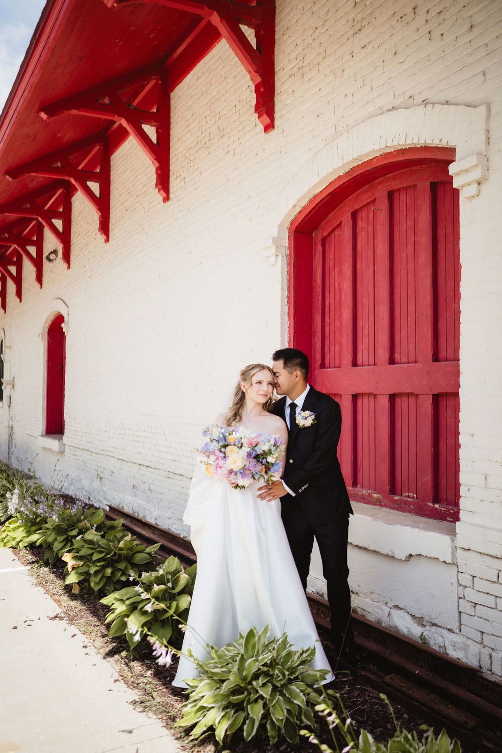 A couple in wedding attire poses for romantic photos against a white wall with red doors and green foliage.