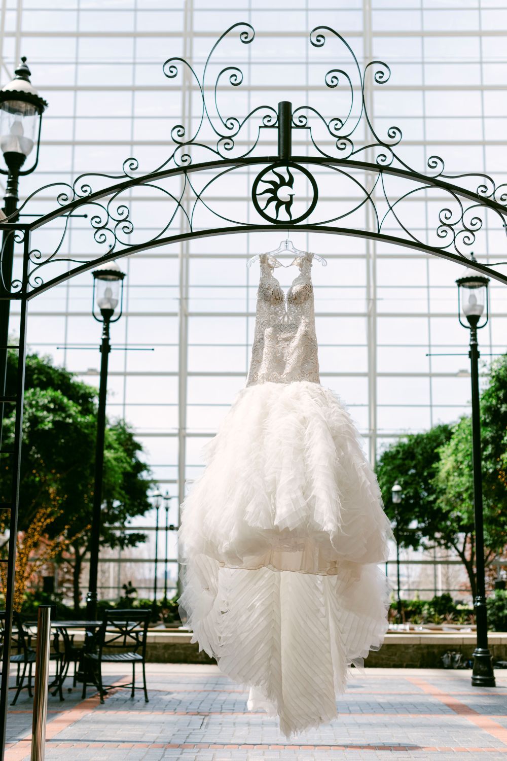 White wedding dresses hanging from decorative iron arches against a bright window backdrop.