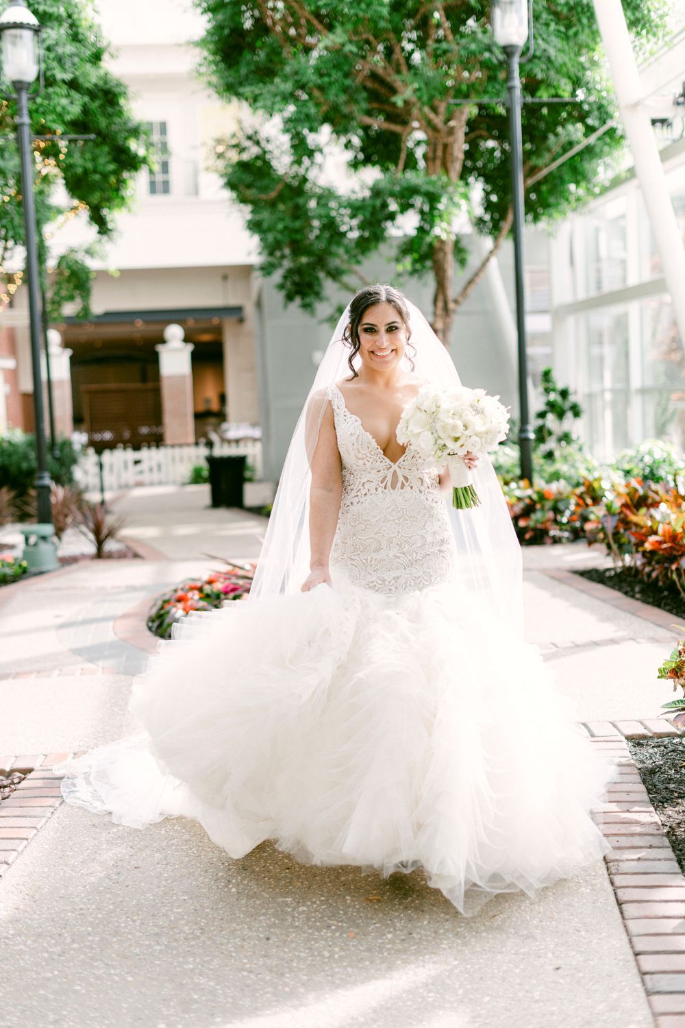 A bride in a flowing white gown poses in an outdoor garden setting with blooming flowers.