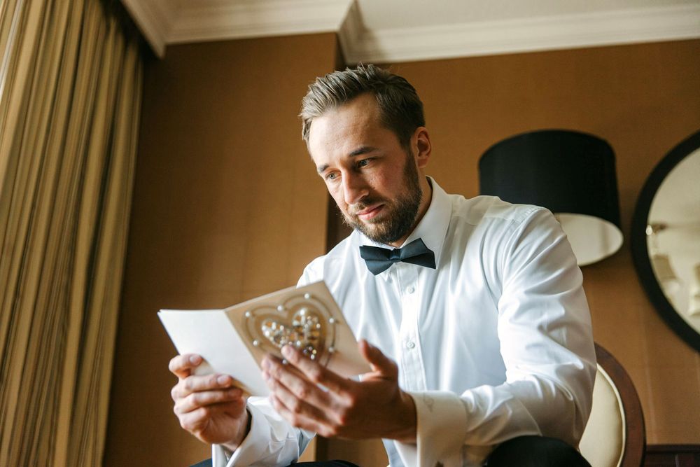 Person in white dress shirt and bow tie reads from paper document in elegant room with clock on wall.