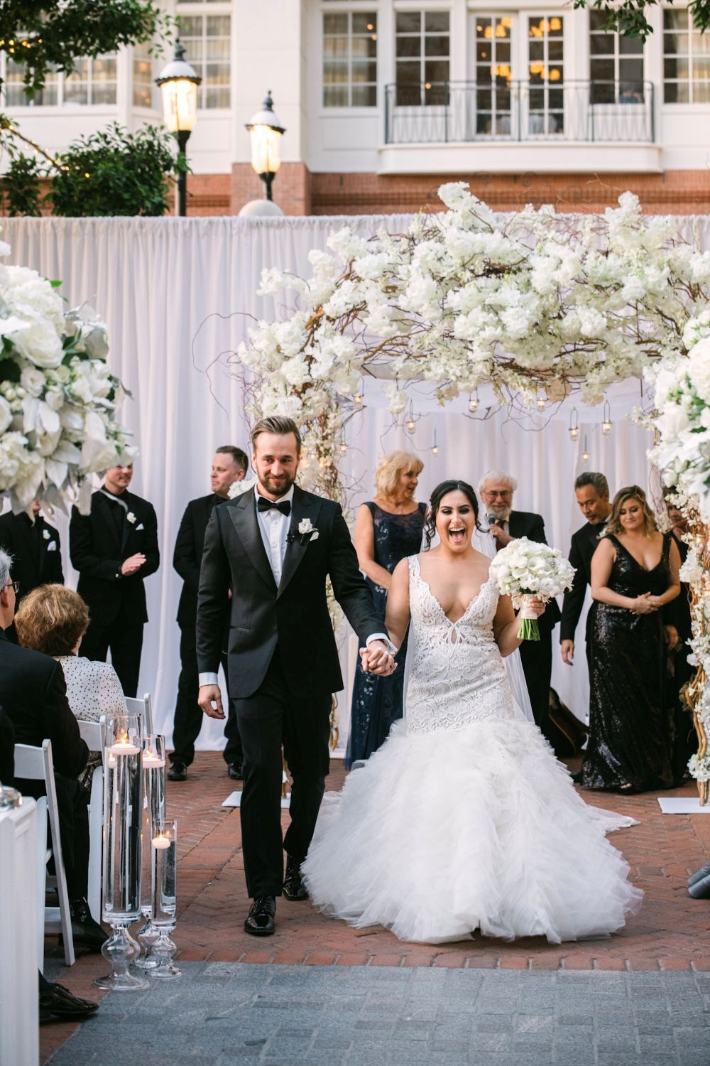 Wedding party gathers around elegant floral arch during outdoor ceremony with white decor and formal attire.