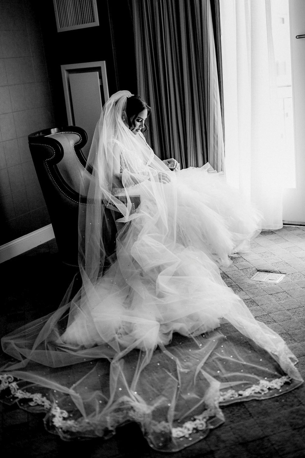 A flowing white wedding veil billows and cascades across a carpeted floor in a dramatic black and white photograph.