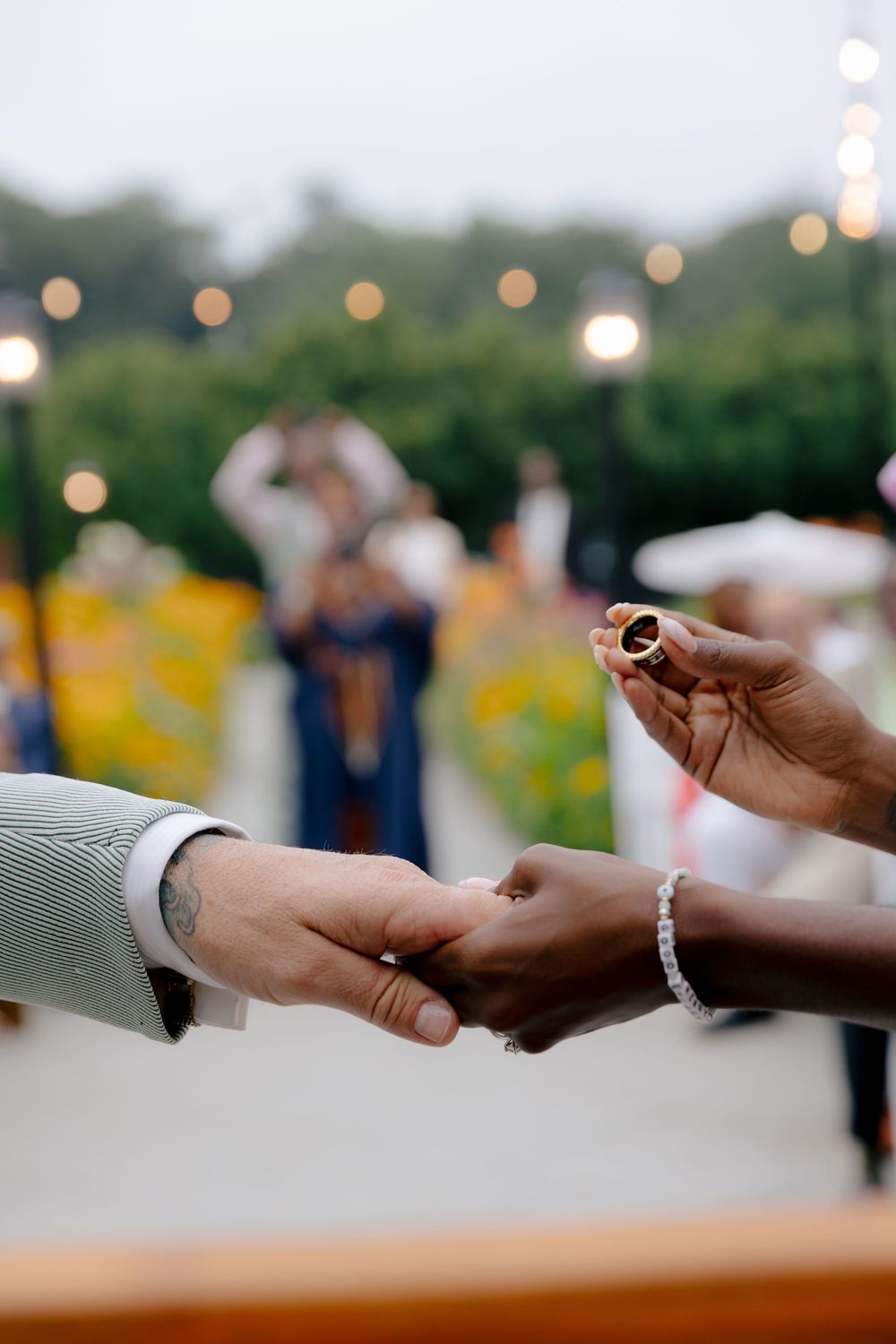Close-up of hands exchanging wedding rings during outdoor ceremony with string lights and sunflowers in background.
