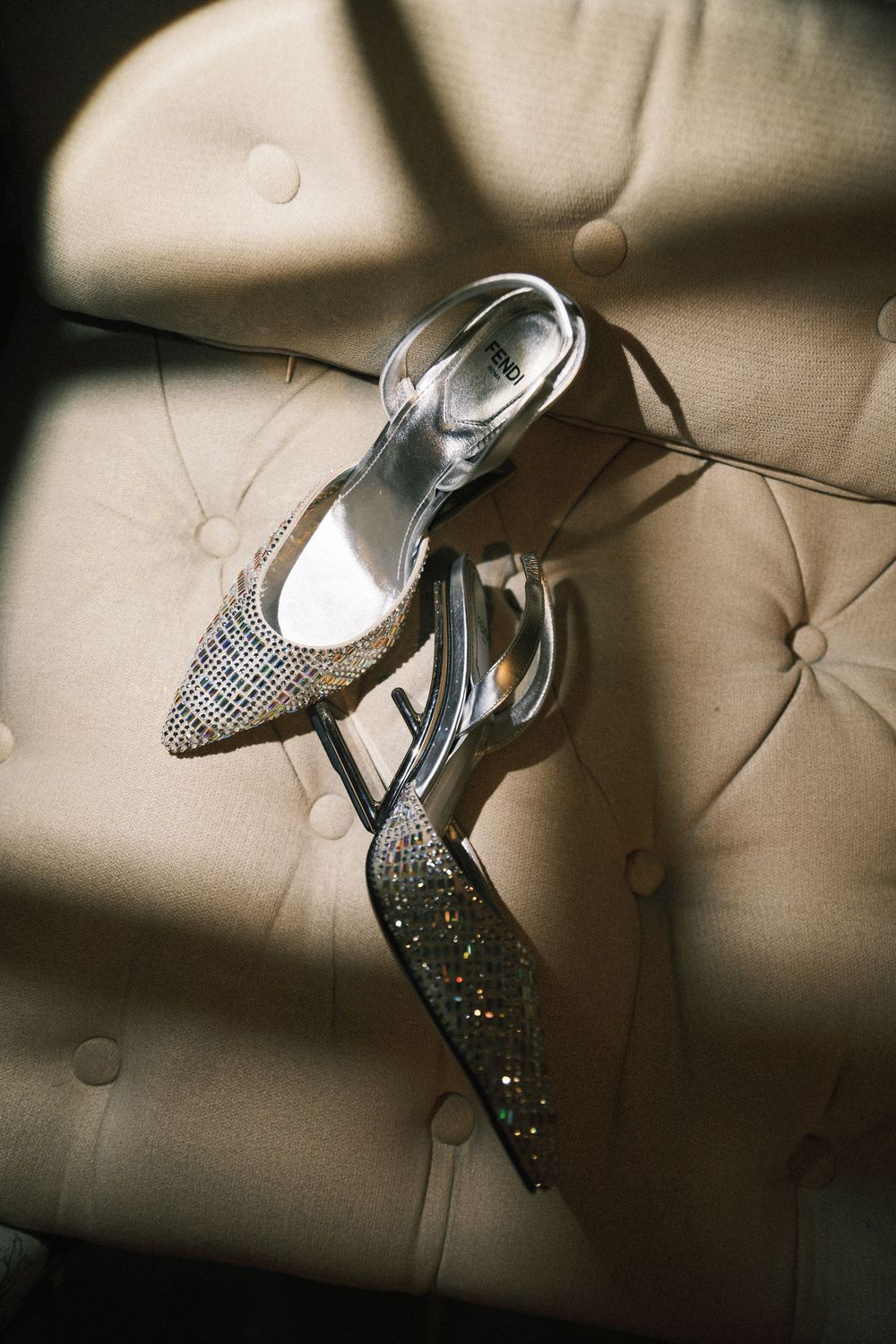 Sparkly silver pointed-toe high heel shoes displayed on a beige tufted leather surface.