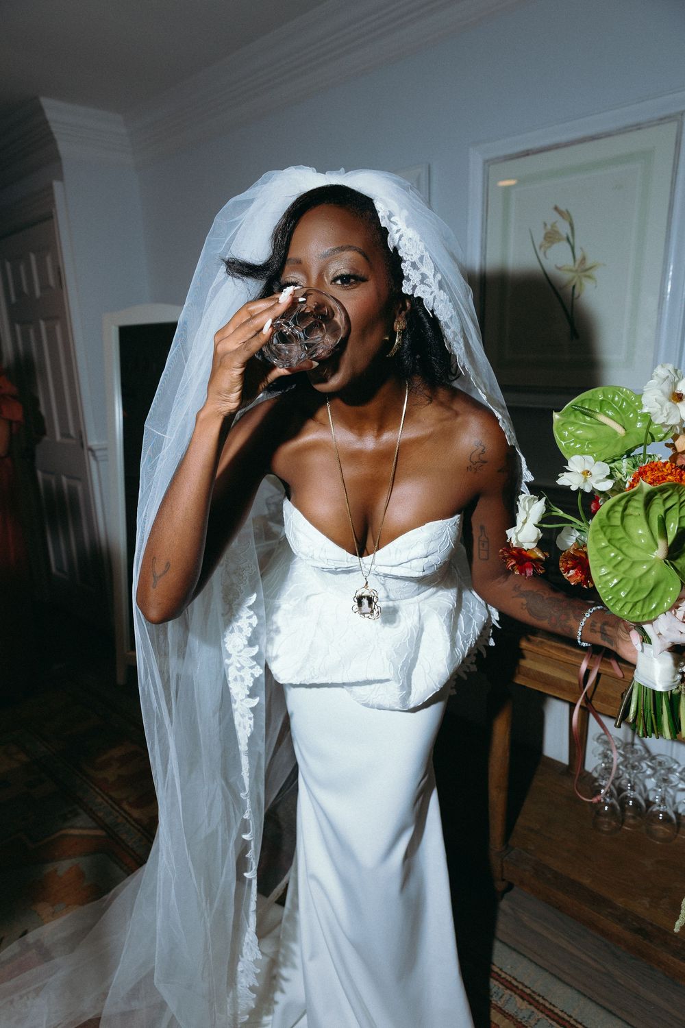 A bride in a white wedding dress and veil takes a celebratory drink during wedding preparations.