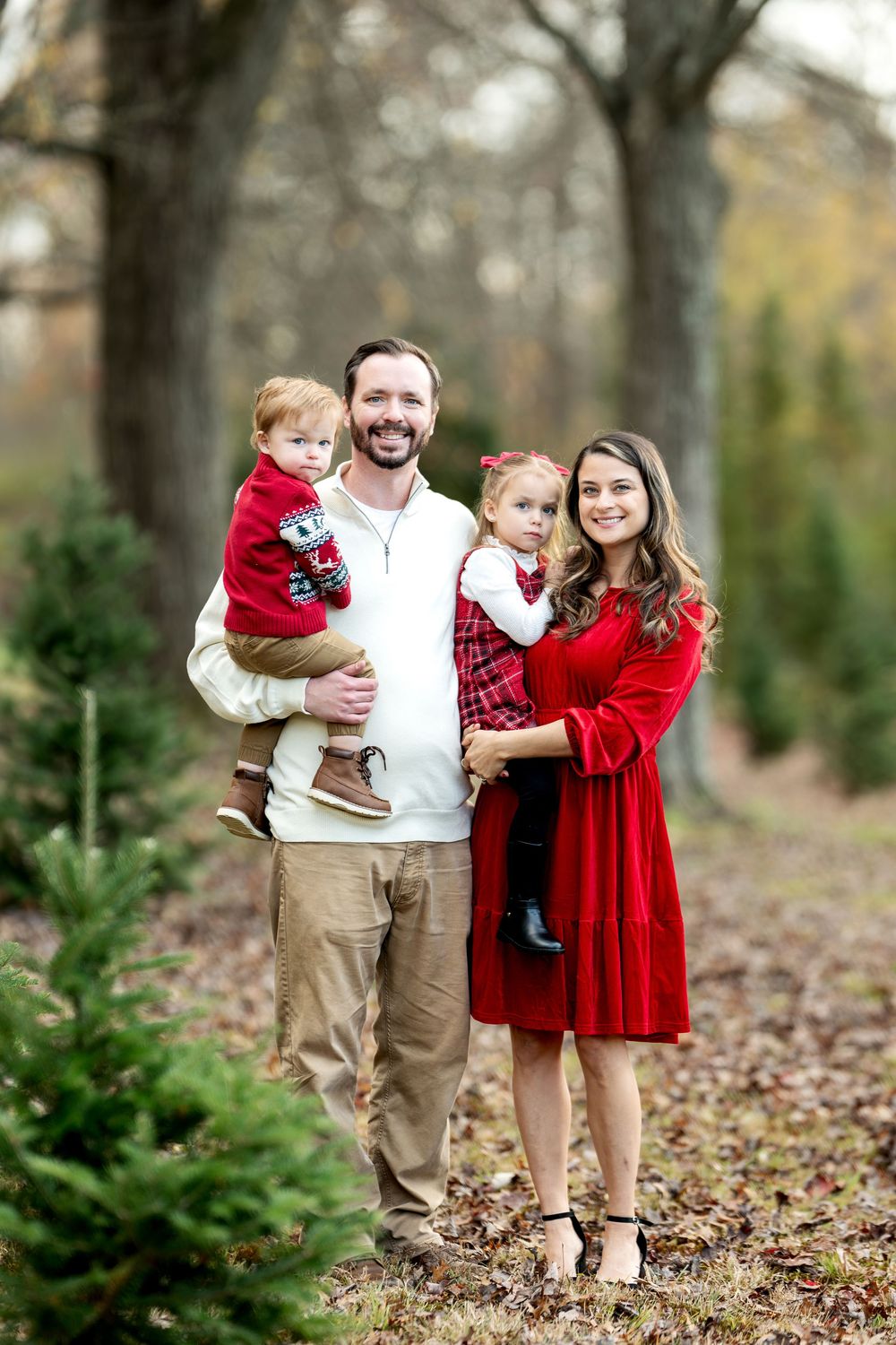 A cheerful family of four poses among Christmas trees, dressed in festive red outfits.